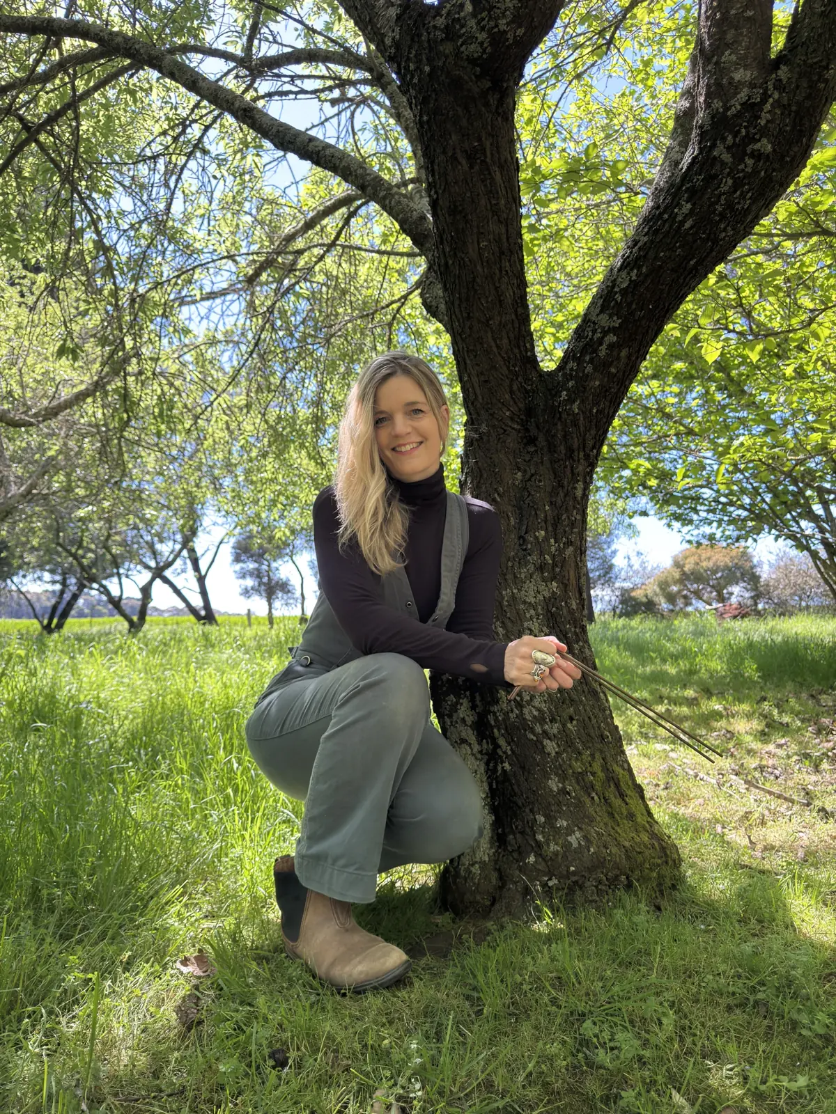Kali smiling beside a tree with her dowsing rods